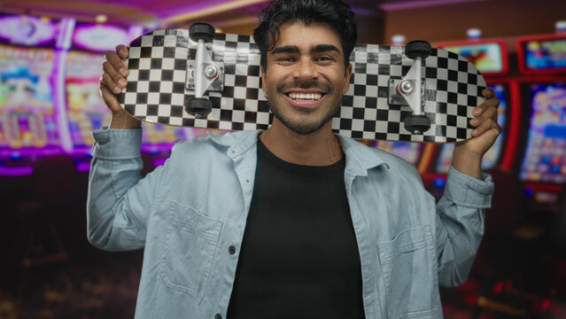 Young hispanic man holds skateboard behind shoulders and smiles broadly near slot machines in casino; playfulness.