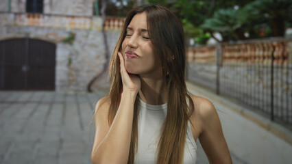Woman holds cheek with hand, closes eyes in front of stone building on street in daylight; reflection.