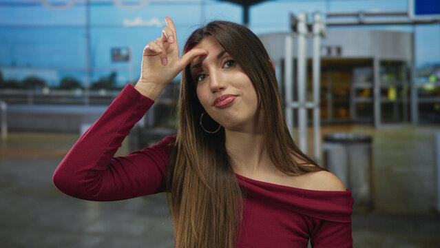 Young hispanic woman with burgundy top makes loser gesture to forehead with hand in airport terminal; mockery.