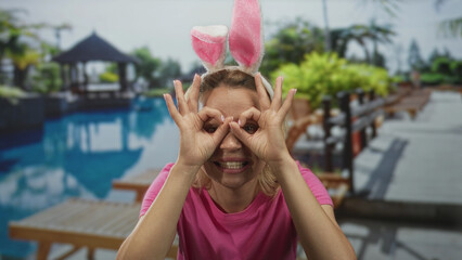 Young blonde woman wearing fluffy bunny ears headband makes ok sign beside resort building poolside...