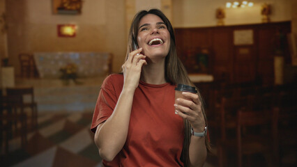 Young hispanic woman in church talking on phone and enjoying coffee indoors, conveying a relaxed and joyful atmosphere amidst religious setting.