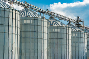 Several metallic grain silos with cone shaped tops stand in a row. Overhead, a steel conveyor system connects them on a partly cloudy day