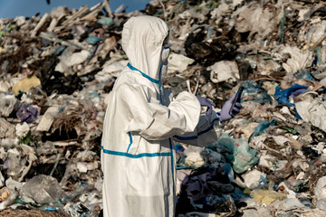 A person wearing a white hazmat suit and blue gloves uses a smart phone in front of a large pile of trash at a landfill, apparently analyzing waste