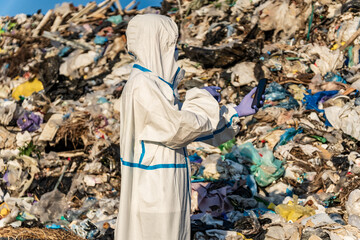 At a landfill, a person wearing a white protective suit and purple gloves examines a mobile phone. A mountain of garbage is in the background