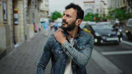 Hispanic man with a beard wearing denim jacket on a busy street, reflecting outdoors, possibly experiencing shoulder pain amidst urban traffic scenery.