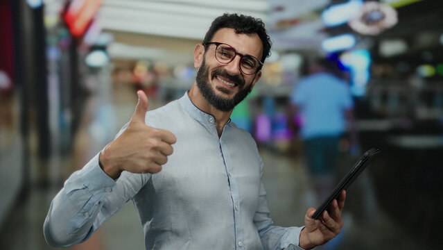 Hispanic man with beard smiling in a shopping mall while holding a tablet, engaging with technology in a vibrant retail environment.
