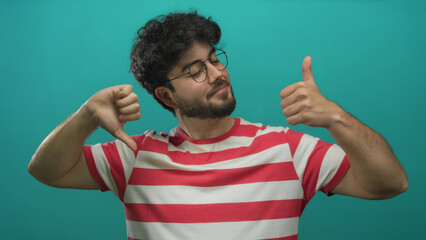 Young man with beard in striped shirt showing thumbs up and thumbs down against green background,...