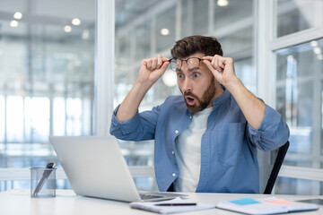 A man in an office looks at a laptop with great surprise, adjusting his glasses.