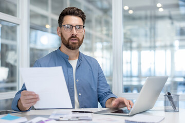 Fototapeta premium A business professional working on a laptop while holding a document, sitting at an office desk with a modern glass-walled backdrop.