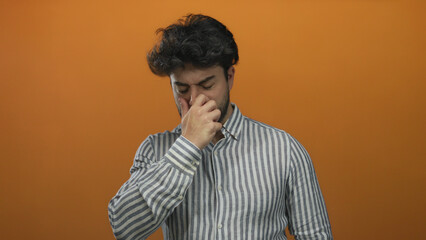 Obraz premium Young hispanic man with a beard covering his nose from a bad smell against an orange background, showing a disgusted expression and wearing a striped shirt.