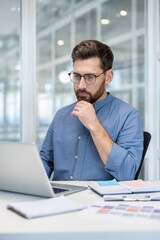 Fototapeta premium A professional individual works on a laptop in a modern, bright office environment. They appear thoughtful, concentrated, and engaged with their tasks, surrounded by organized office essentials.