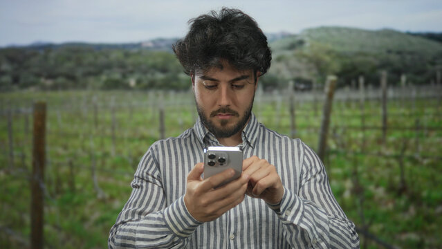 Young man with beard using smartphone outdoors in a park-like setting, surrounded by lush greenery and a vineyard backdrop under a cloudy sky, portraying a relaxed outdoor experience.