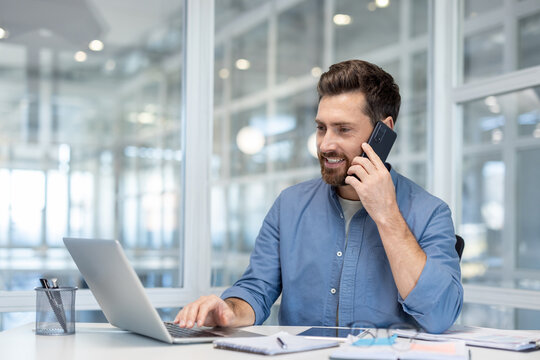 Business professional talking on smartphone while working on a laptop in a bright office setting. Organized desk with documents creates a productive workspace.