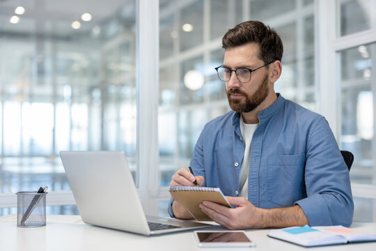 A man wearing glasses, using a laptop and taking notes while sitting in an office. The setting features a well-lit, modern design, promoting an atmosphere of concentration