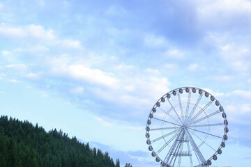 Fototapeta premium White Ferris wheel rising above lush green forest with mountain backdrop, scenic summer view, travel and leisure destination, eco tourism. Ferris wheel stands tall against a deep blue sky