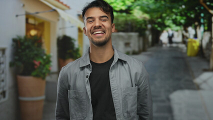 Fototapeta premium Young hispanic man smiling outdoors on a city street, exuding positivity and confidence, while giving a thumbs-up gesture under dappled sunlight amidst urban surroundings.