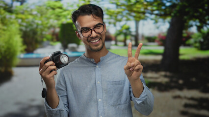 Young hispanic man smiling at camera in a park holding a camera with a peace sign, surrounded by vibrant green nature during daylight.
