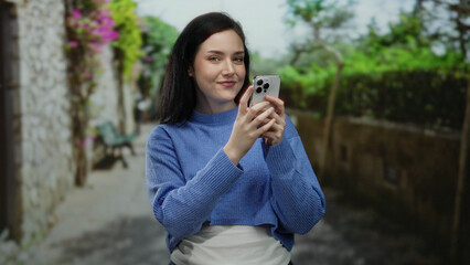 Woman smiling holding smartphone on picturesque street in old town with stone walls and vibrant flowers, capturing a candid moment of urban exploration and connection.