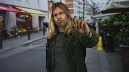 Man with long blond hair wearing a green corduroy jacket crossing arms in x gesture on a busy...