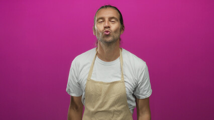 Man with long blond hair and white shirt wearing an apron pointing both hands upward in a pink studio; optimism.