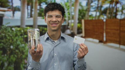 Man holds cash jar filled with bills and blank card with both hands outside building entrance;...