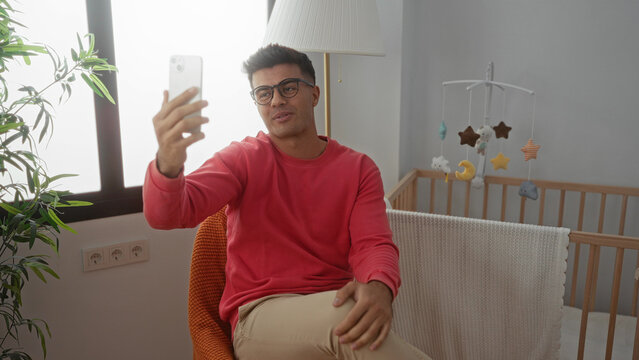 Young man in glasses takes a selfie indoors in a cozy bedroom with a baby crib and a tall plant by the window, sitting comfortably in an orange chair, smiling with relaxed posture.