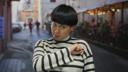 Young chinese woman on restaurant terrace making a brotherhood gesture with her fist on chest, wearing striped sweater outdoors.