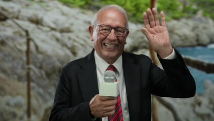 Senior man in suit holding microphone smiling and waving at seaside beach backdrop outdoors, embodying business and media reporting ambiance with elegant gesture.