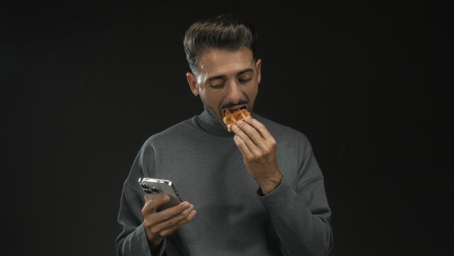 Man eating waffle while holding smartphone in black studio setting; multitasking convenience relaxation. - Powered by Adobe