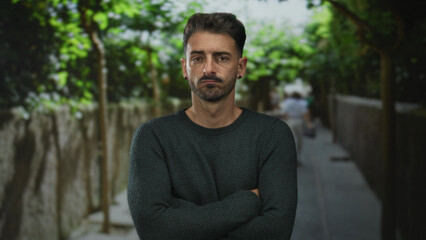 Fototapeta premium Man wearing green sweater with arms crossed on a sunlit street lined with trees and stone walls in a city lane; rejection.