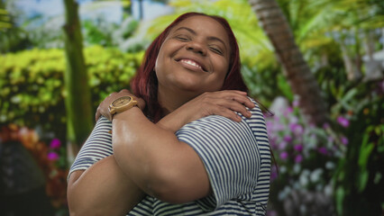 Smiling woman wearing wooden watch hugging arms across chest amid bright green forest; pure self...