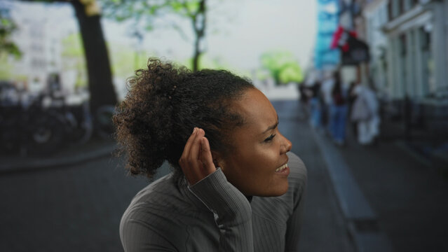 Smiling young african american woman wearing gray zip sweater holds hand to ear gesture in urban street; curiosity.
