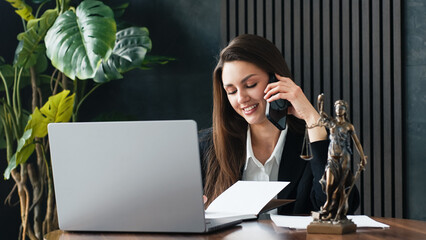 Female lawyer engaged in a phone conversation while working at her desk, with a laptop and legal statue, surrounded by greenery, showcasing a professional and modern office environment
