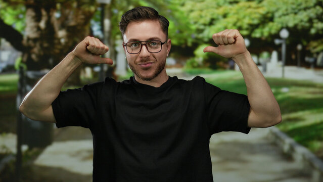 Young man outdoors in park, wearing black shirt, smiling and pointing at himself with thumbs, with trees and pathway in background, showing playful confident attitude.