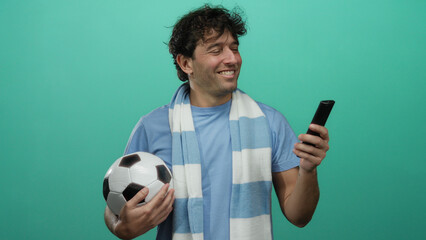 Hispanic man holding football and remote on green background, smiling enthusiastically, capturing the essence of sports fandom and leisure.