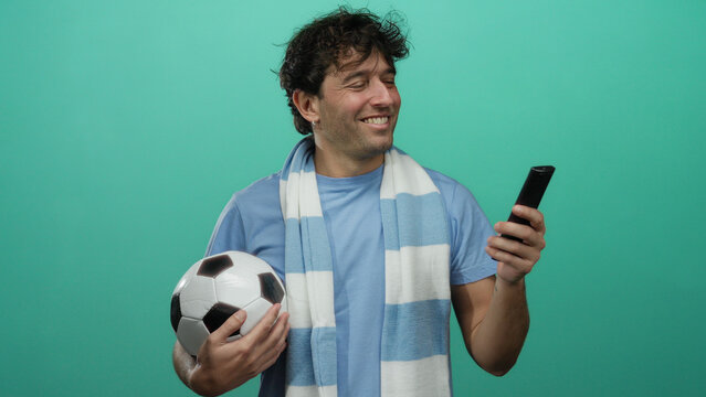 Hispanic man holding football and remote on green background, smiling enthusiastically, capturing the essence of sports fandom and leisure.