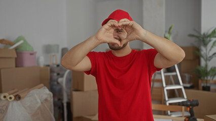 Young hispanic delivery man wearing red cap and red shirt forms heart with hands in new home building; warmth love.