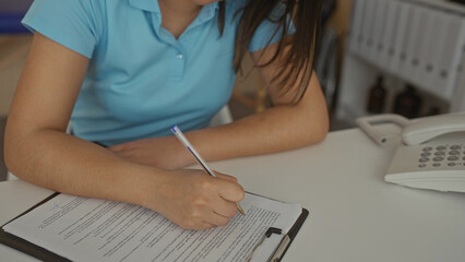 Young woman writing in a clinic workplace, focused on completing forms with a pen in an indoor...