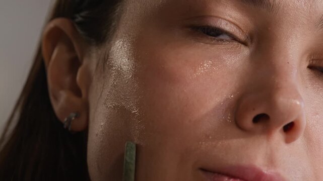 Close-up of young brunette woman with brown eyes standing in front of mirror during self care ritual and using gua sha scraper for facial massage with serum