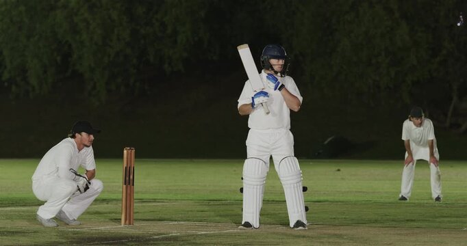 Bowler delivering ball, male batter playing block shot defending wicket on pitch under floodlights