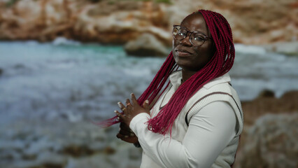 Woman smiling in front of a scenic seaside with vibrant pink braids and glasses, capturing a joyful...