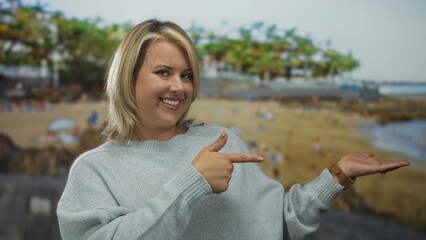 Woman smiles and makes thumbs up gesture and holds open palm to side presenting space in studio with blurred beach view on screen; approval.