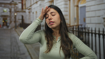 Fototapeta premium Woman outdoors on street with contemplative expression, touching forehead and chin, suggesting thoughtfulness in urban environment, with buildings and iron fence visible.