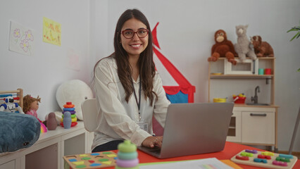 Young hispanic woman teacher working indoors on a laptop in a kindergarten surrounded by educational playful toys and drawings, creating an engaging preschool learning environment.