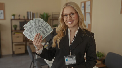 Woman in office holding dollar bills with a confident smile, wearing glasses and business attire, representing financial success and stability in a professional indoor environment.