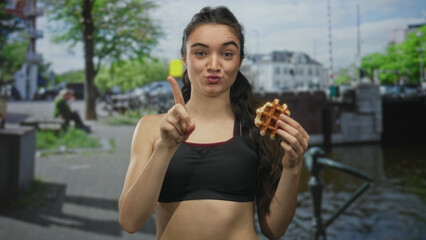 Fototapeta premium Hispanic teenage woman holds waffle and points finger in refusal on street by canal railing with playful pout; denial.