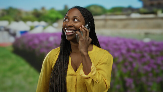 Woman joyfully talking on smartphone in vibrant yellow shirt, standing outdoors in a sunny park with purple flowers and greenery in the background, exuding happiness and excitement. - Powered by Adobe