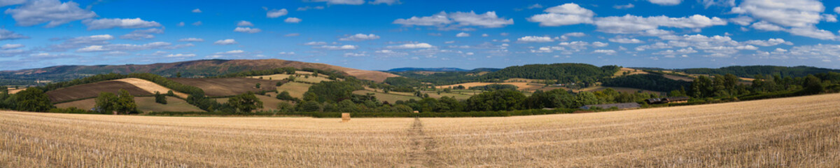 Late-summer panorama of a harvested field with a single straw bale, rolling patchwork hills and scattered clouds in Shropshire, UK.