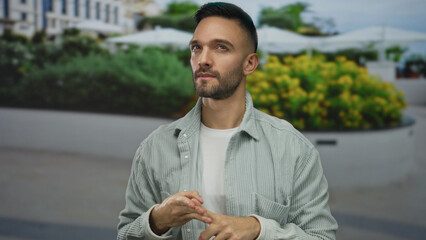 Hispanic young man counts with fingers on city street surrounded by urban scenery with vibrant flowers, conveying thoughtfulness and concentration in a bustling outdoor environment.