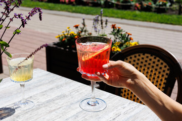 Hand holding red summer cocktail with orange slice outdoors, Close-up of a hand holding a red cocktail with orange slice and ice on a sunny outdoor terrace surrounded by flowers and greenery.  